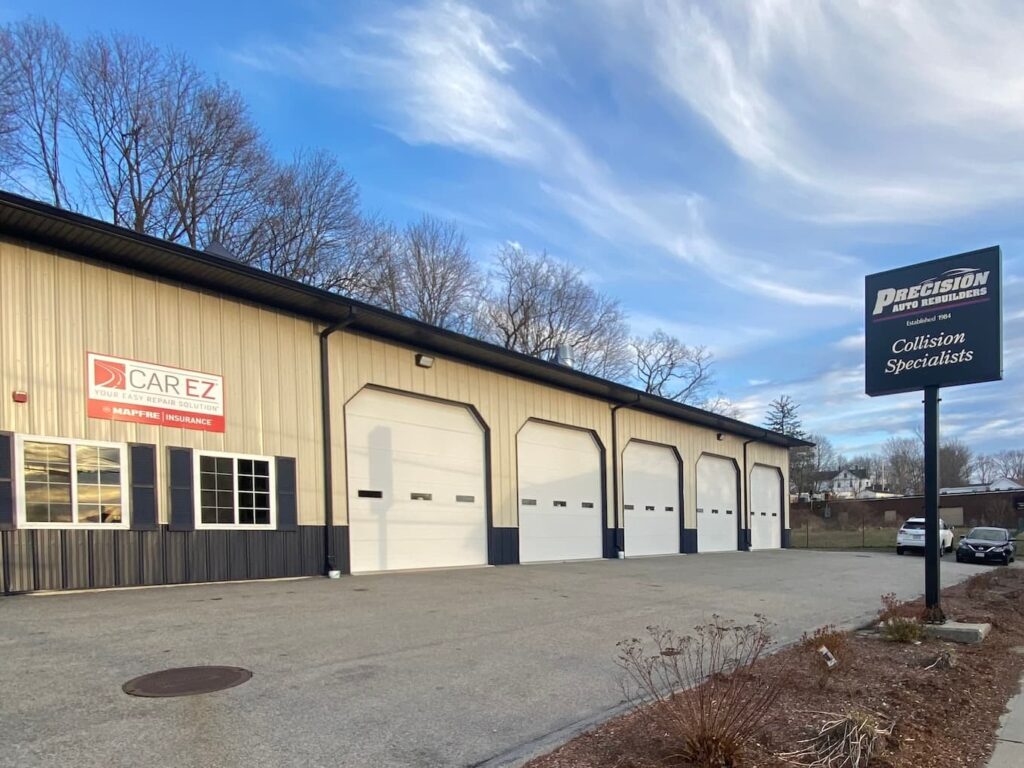 A large blue building with multiple garage doors, a Carez insurance logo, and a Precision Auto Specialists sign, surrounded by trees and a paved parking area.